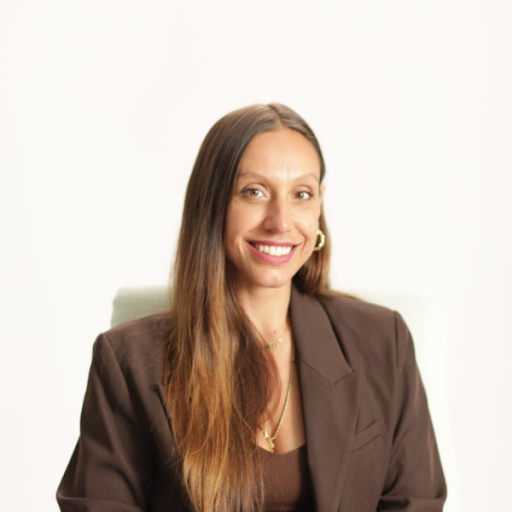 Person with long brown hair and a brown blazer smiling against a plain white background.